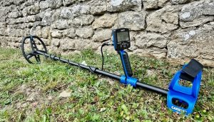 Le détecteur de métaux Garrett Vortex VX5 est positionné contre un mur de pierres anciennes et rustiques, qui sert de toile de fond texturée. L'appareil, avec son design moderne et ses composants noirs et bleus, crée un contraste intéressant avec la pierre patinée par le temps. Le détecteur est prêt à l'emploi, comme s'il attendait sa prochaine aventure de prospection.